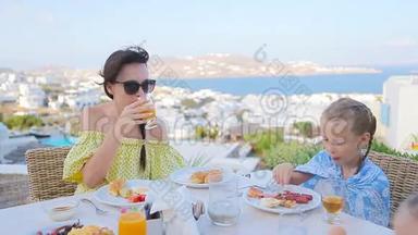 一家人在室外咖啡馆吃早餐，在Mykonos镇有令人惊叹的景色。 可爱的女孩和妈妈喝着新鲜果汁